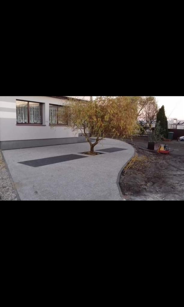 Szara, przepuszczalna nawierzchnia z żywicy epoksydowej ułożona wokół drzewa przed domem, z ciemnymi prostokątnymi wstawkami i ubijarką w tle.
