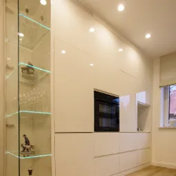 Nowoczesna, biała zabudowa kuchenna z połyskiem, z wbudowanym piekarnikiem i przeszkloną witryną z podświetlanymi półkami, na których stoją kieliszki i ozdobne figurki.