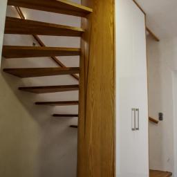 Usługi Stolarskie Andrzej Prabucki - Spiralne schody drewniane zintegrowane z szafą wnękową z białymi frontami i podłużnymi uchwytami, oświetlenie punktowe na ścianie.