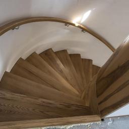 Usługi Stolarskie Andrzej Prabucki - Spiralne schody drewniane z jasnego drewna, widok z góry, z delikatnym oświetleniem padającym z góry.