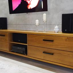 Usługi Stolarskie Andrzej Prabucki - Drewniana szafka RTV z szufladami i półkami, na której stoją głośniki, świeczki w szklanych świecznikach i porcelanowa cukiernica, na tle ściany z betonowych płyt i telewizora.