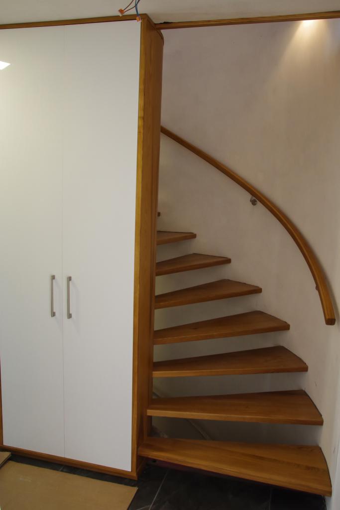 Biała szafa w zabudowie obok drewnianych, kręconych schodów z poręczą, oświetlonych punktowym światłem.