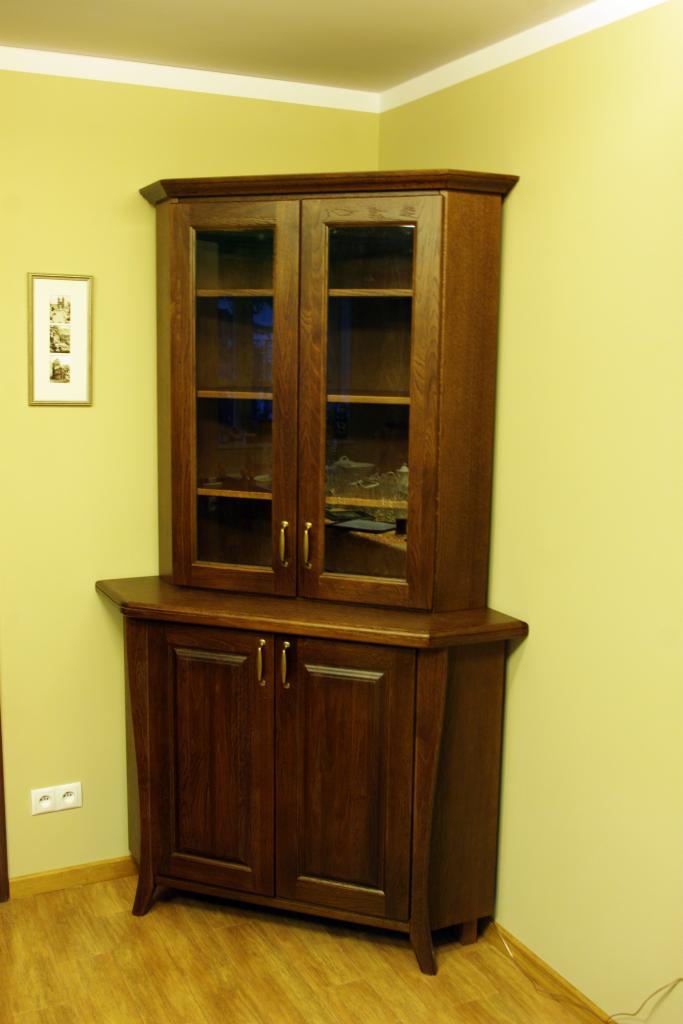 Ciemnobrązowa, narożna witryna z przeszkloną górną częścią i dolną szafką, ustawiona w rogu pokoju z żółtymi ścianami i białym sufitem.