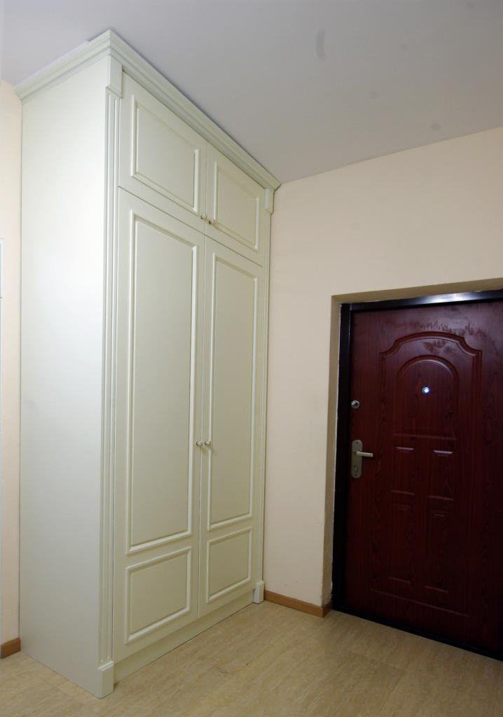 Biała szafa dwudrzwiowa z nadstawką, w klasycznym stylu, z dekoracyjnymi frezowaniami na frontach, ustawiona w przedpokoju obok ciemnych drzwi wejściowych.