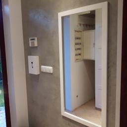 Remonty - Wnętrze przedpokoju z szarą ścianą, na której wisi białe lustro w ramie, odbijające otwartą skrzynkę z bezpiecznikami; obok drzwi wejściowe z widokiem na ogród.