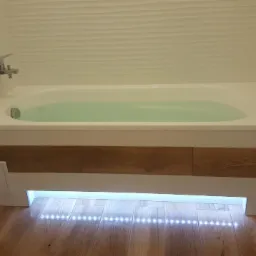 Nowoczesna wanna z obudową z drewnopodobnych paneli, podświetlona listwą LED, na tle białej ściany z dekoracyjną fakturą i drewnianej podłogi.