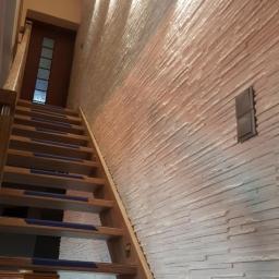 Remonty - Drewniane schody z niebieskimi nakładkami antypoślizgowymi prowadzące na górę, oświetlone kinkietami na ścianie wykończonej dekoracyjną strukturą imitującą kamień.