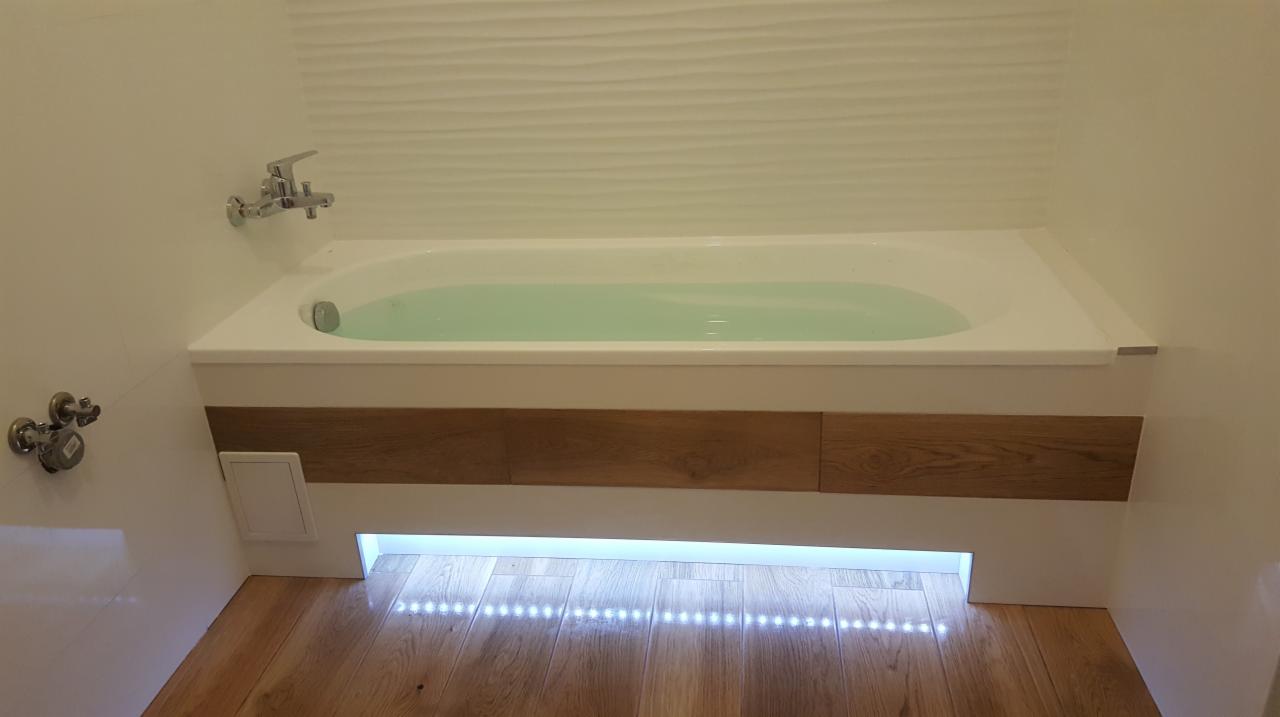 Nowoczesna wanna z obudową z drewnopodobnych paneli, podświetlona listwą LED, na tle białej ściany z dekoracyjną fakturą i drewnianej podłogi.