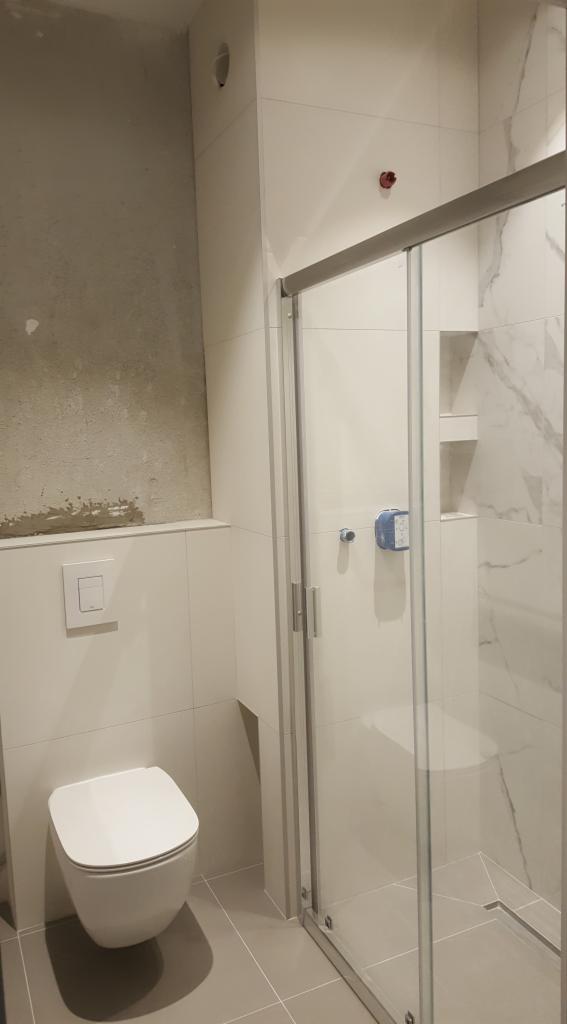 Nowoczesna łazienka z podwieszaną toaletą, kabiną prysznicową z przesuwnymi drzwiami i fragmentem ściany przed wykończeniem, ukazującym surowy tynk.