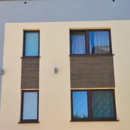 Fragment elewacji budynku po remoncie z beżową i szarą farbą, brązowe okna i imitacja drewnianej okładziny.