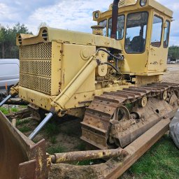 firma usługowa - Żółty, używany spychacz gąsienicowy z widocznymi śladami rdzy i błota, zaparkowany na trawiastym terenie z widocznym lasem w tle i fragmentem białego busa po lewej stronie.