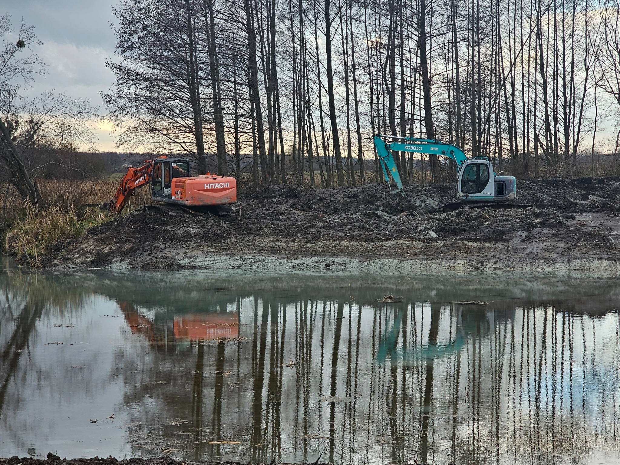 Dwie koparki, pomarańczowa Hitachi i turkusowa Kobelco, pracują na błotnistym brzegu zbiornika wodnego, ich odbicia widoczne w wodzie, w tle rząd wysokich, nagich drzew.