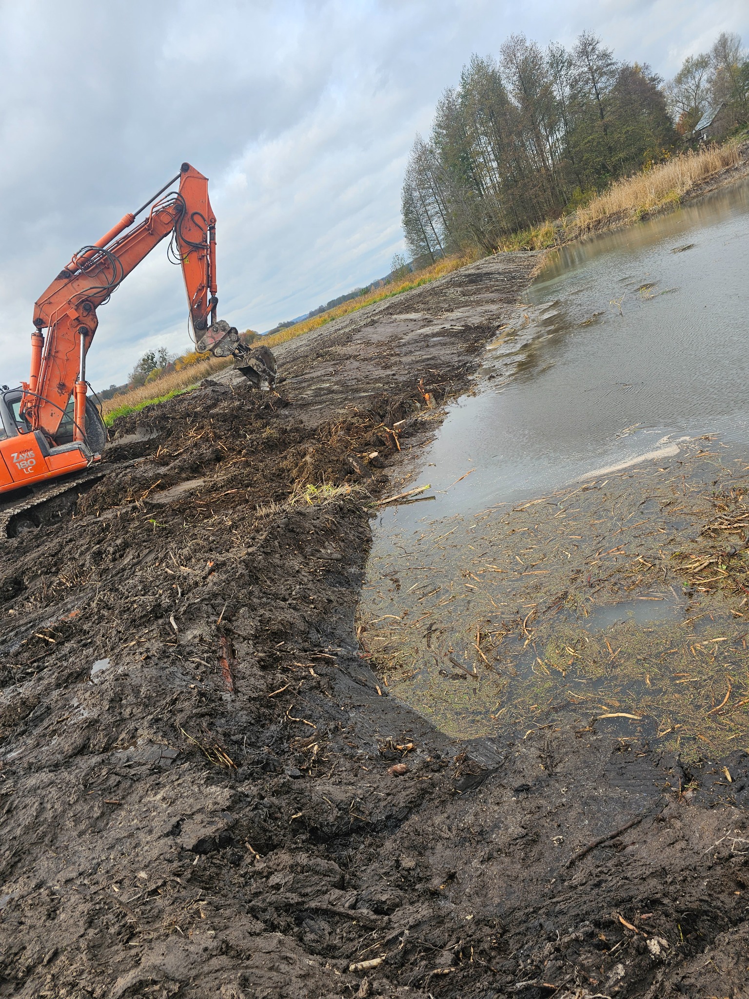 Pomarańczowa koparka Hitachi AXIS 180 LC na brzegu zbiornika wodnego, świeżo wykopana ziemia i roślinność w wodzie, pochmurne niebo w tle.