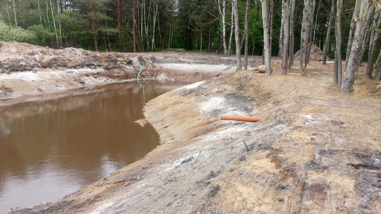 Wykopany staw w lesie, brudna woda, widoczne brzegi z piasku i gliny, rura odpływowa na brzegu, w tle drzewa.