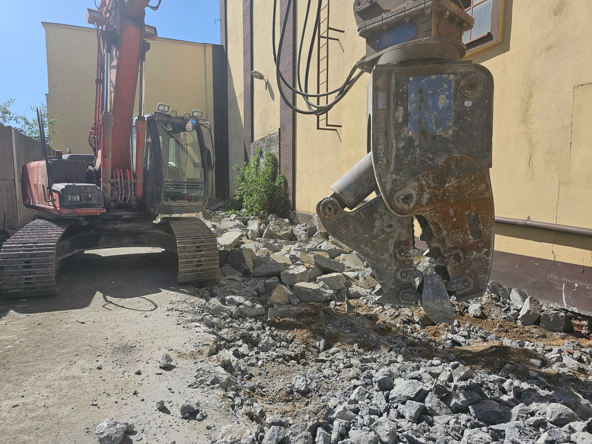 Czerwony, gąsienicowy koparka z hydraulicznymi nożycami do betonu kruszy gruz na placu budowy, widoczne fragmenty ściany budynku z drabiną.