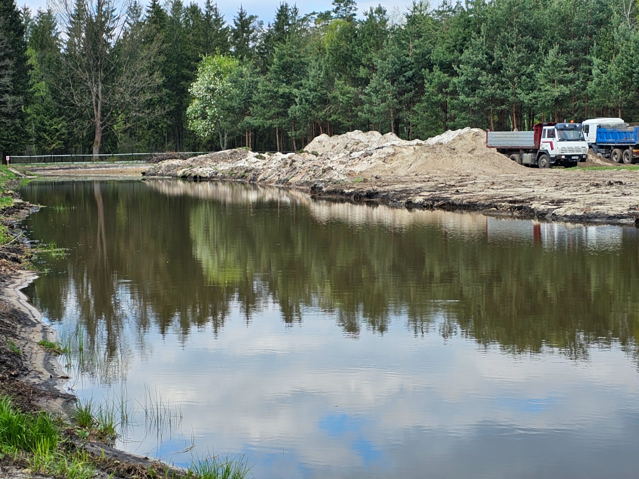 Powierzchnia wykopanego stawu odbija las i niebo; brzeg usypany ziemią, w tle zaparkowane ciężarówki.