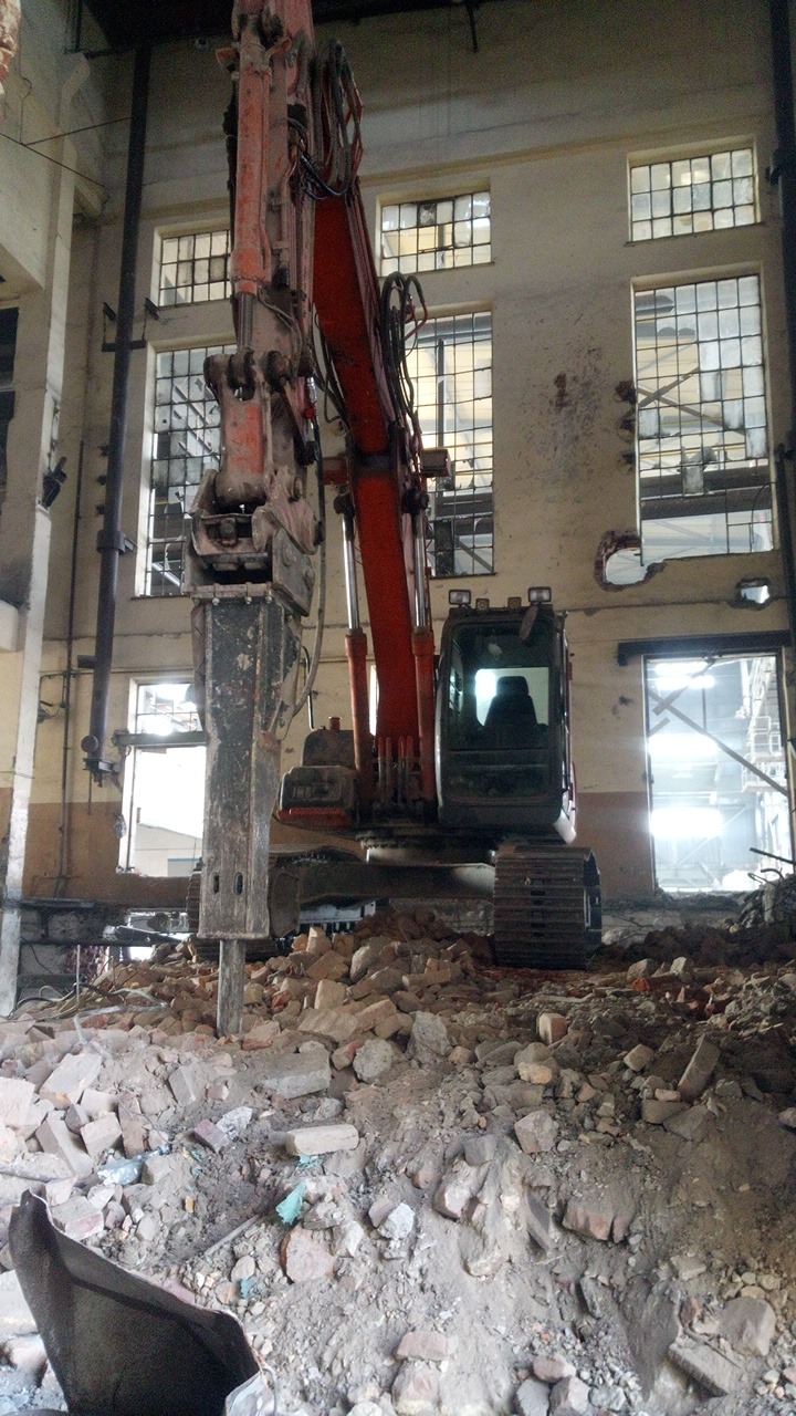 Ciężki hydrauliczny młot udarowy zamontowany na koparce gąsienicowej w trakcie pracy wewnątrz budynku, gruzowisko na pierwszym planie, widoczne okna z kratami w tle