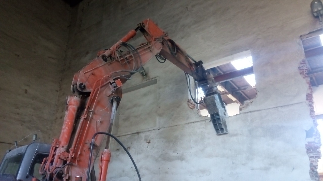 Pomarańczowy młot hydrauliczny zamontowany na koparce wyburza fragment ściany wewnątrz budynku. Fragment dachu z widoczną konstrukcją stalową. Częściowo odsłonięta cegła.