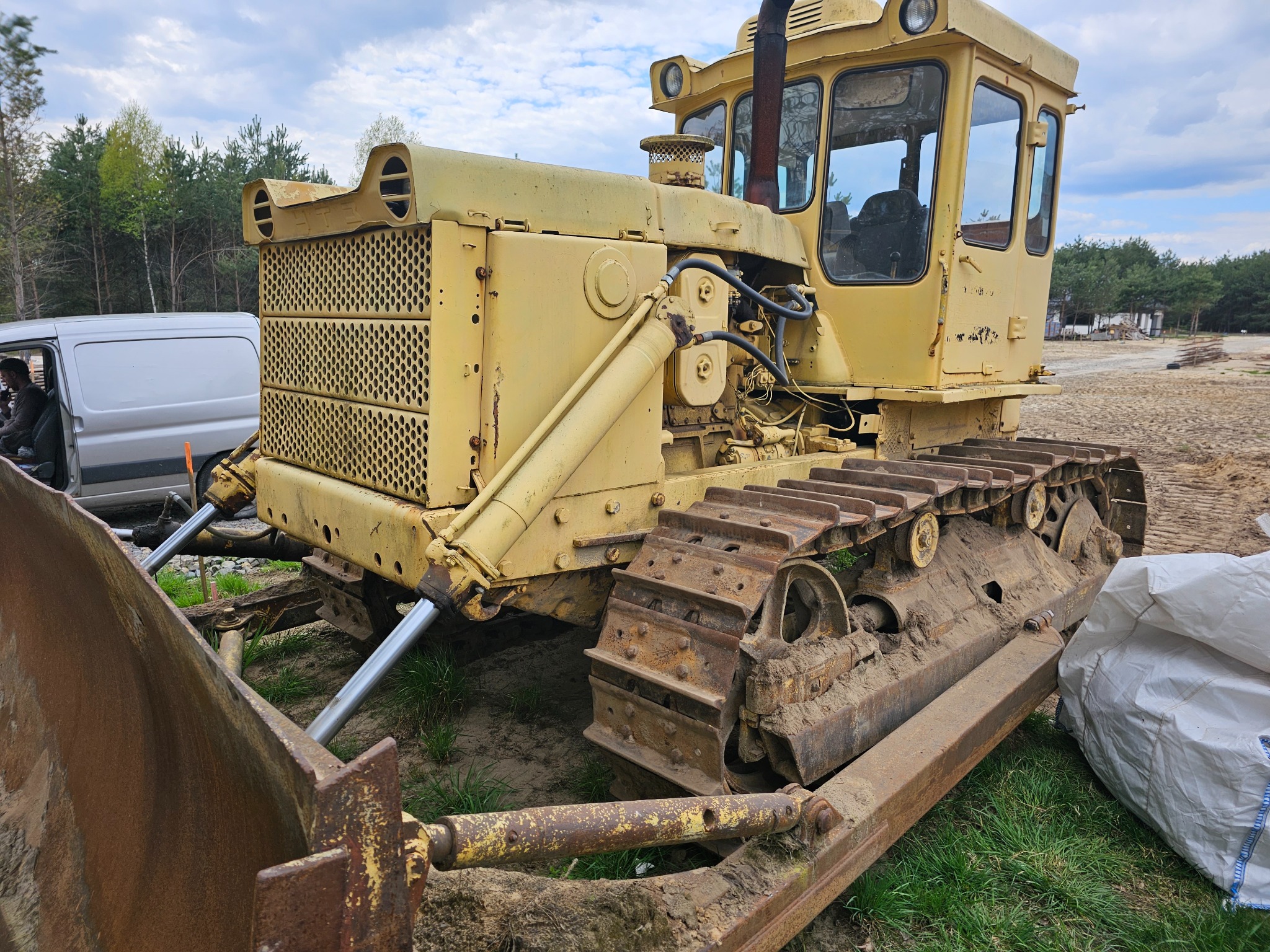 Żółty, używany spychacz gąsienicowy z widocznymi śladami rdzy i błota, zaparkowany na trawiastym terenie z widocznym lasem w tle i fragmentem białego busa po lewej stronie.