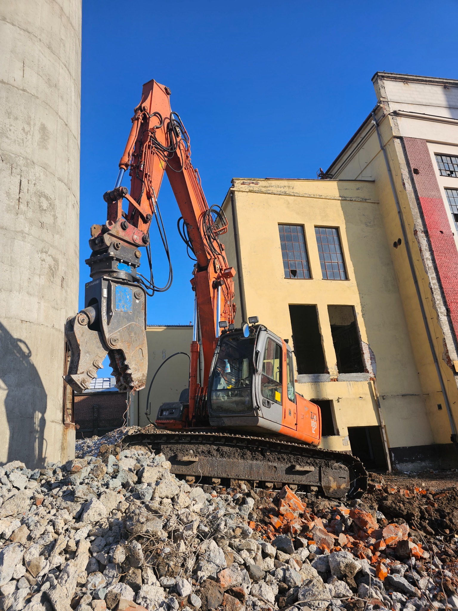 Pomarańczowa koparka z hydraulicznymi nożycami do betonu, rozbiórka budynku z żółtą elewacją i uszkodzonymi oknami, gruzowisko u podstawy, betonowy silos obok, błękitne niebo.