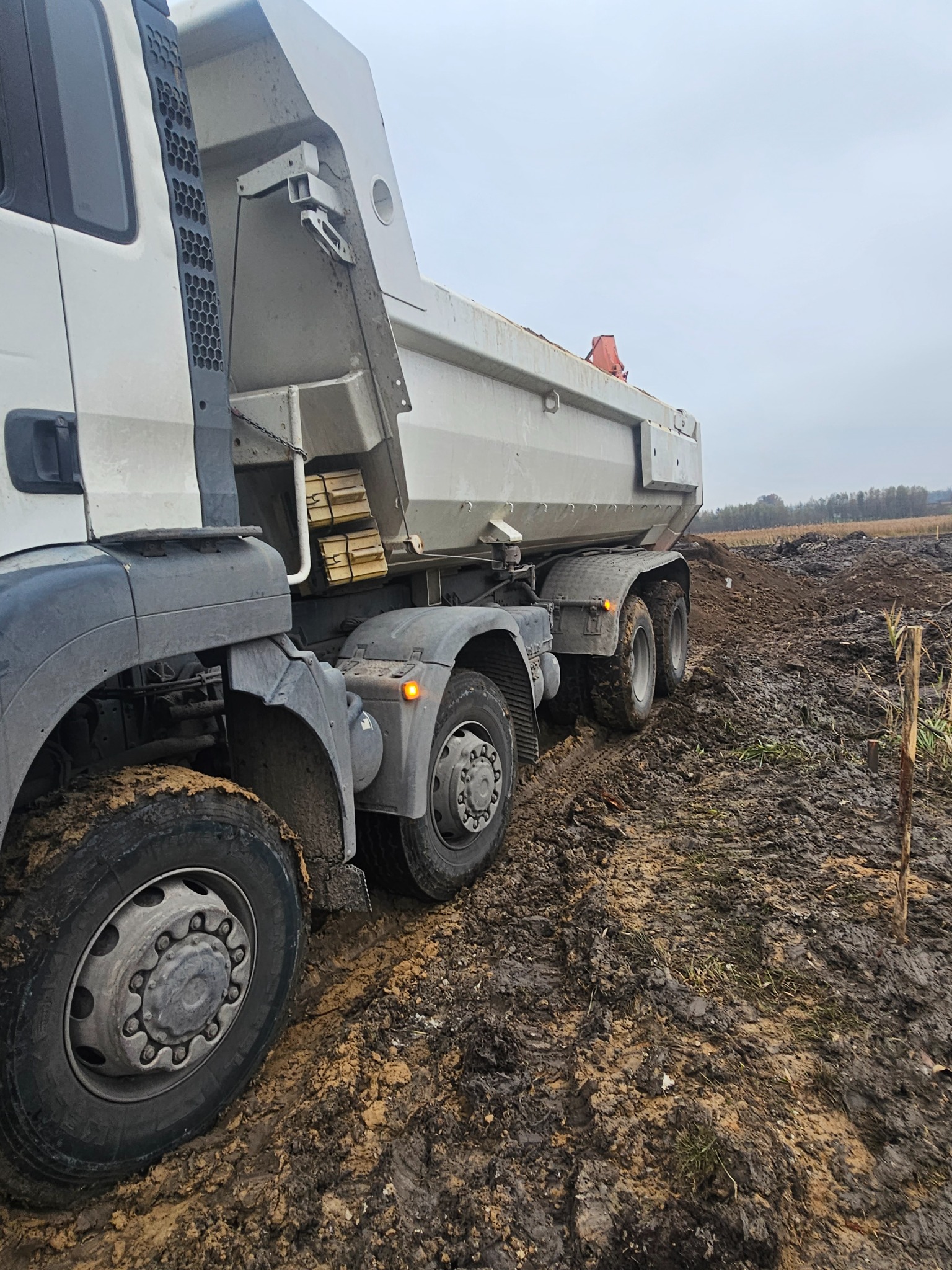 Ciężarówka wywrotka na grząskim, błotnistym terenie budowy, widoczne świeżo wykopane zagłębienie w ziemi i fragment horyzontu.