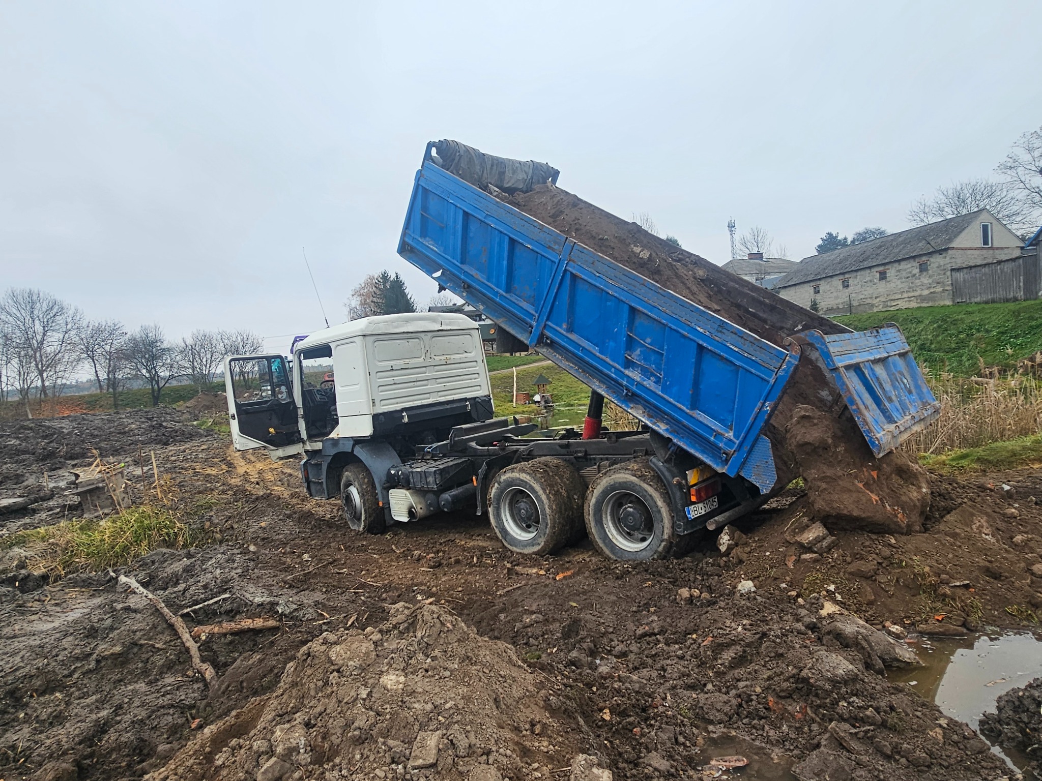 Ciężarówka z podniesioną, niebieską skrzynią ładunkową wysypuje ziemię na błotnisty teren budowy. Otwarta kabina kierowcy, widoczne budynki w tle.
