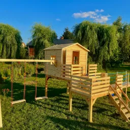 Drewniany domek na palach z huśtawką i zjeżdżalnią, otoczony zielonym trawnikiem i drzewami w słoneczny dzień.