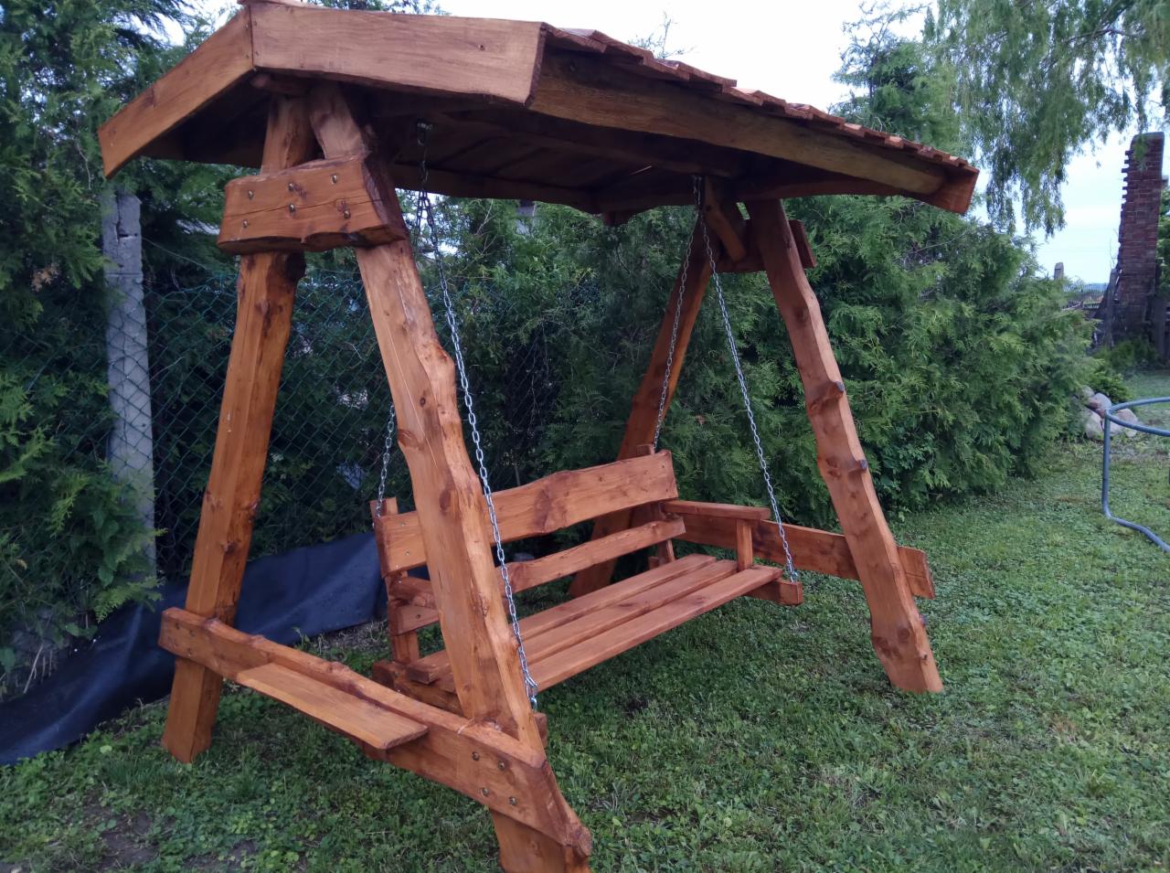 Drewniana huśtawka ogrodowa z zadaszeniem, wykonana z bejcowanego drewna, zawieszona na łańcuchach, widoczna na tle zieleni ogrodowej.
