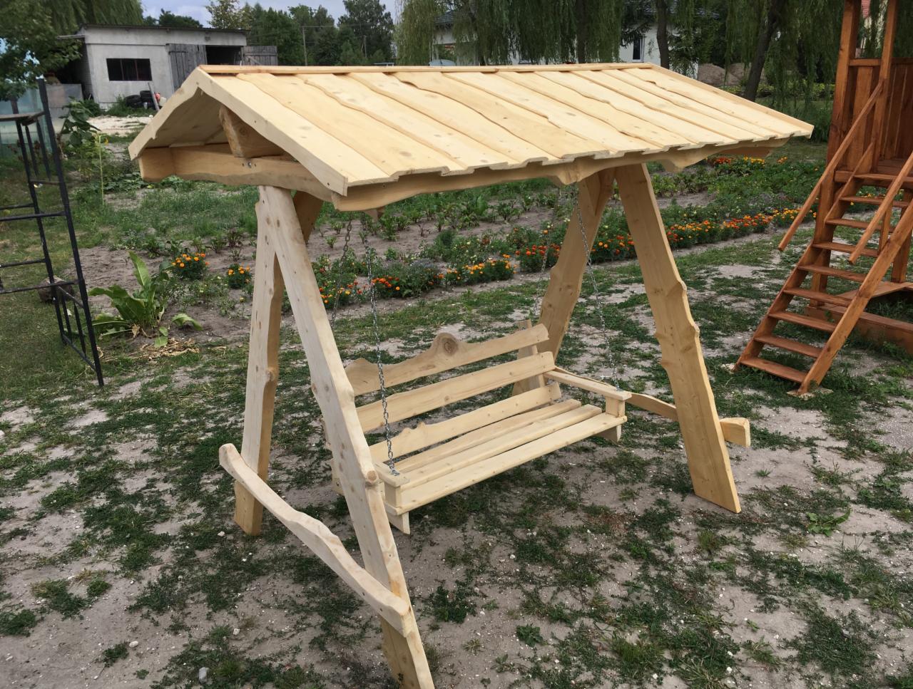 Drewniana huśtawka ogrodowa z zadaszeniem, wykonana z surowego drewna, zawieszona na łańcuchach, na tle trawnika i kwiatów.