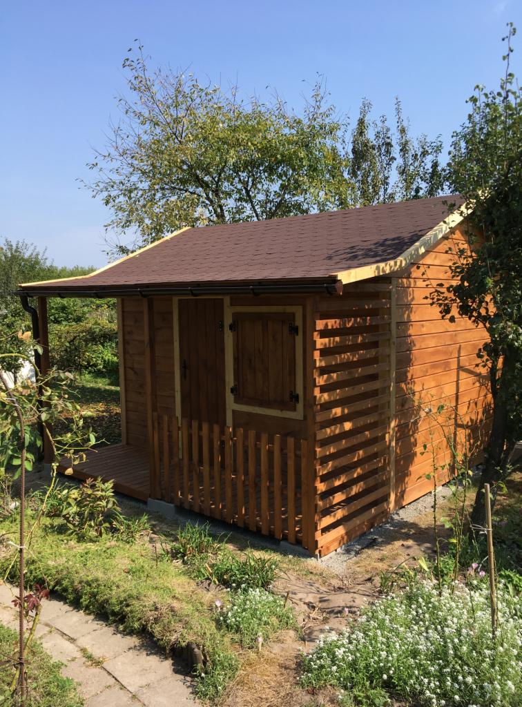 Drewniana konstrukcja z brązowym dachem, prawdopodobnie domek ogrodowy lub mała altana, z drewnianymi okiennicami i małym tarasem, otoczona zielenią i kwiatami.