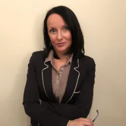 Portret kobiety w czarnej marynarce z białym obszyciem i beżowej koszuli, trzymającej okulary, na jasnym tle.