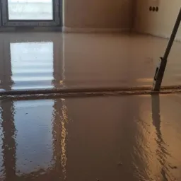 Świeżo wylana, mokra posadzka wyrównywana narzędziem z długim uchwytem, odbicie okna w gładkiej powierzchni.