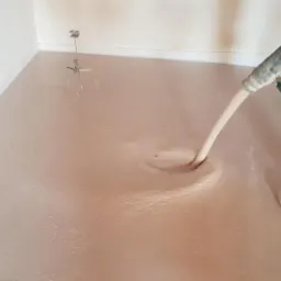 Aplikacja wylewki anhydrytowej: wylewka w trakcie rozprowadzania z węża, widoczne buty robocze i mieszadło na ścianie, w tle biała ściana.