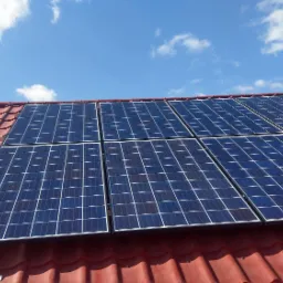 Panele słoneczne zamontowane na czerwonym dachu z dachówki ceramicznej pod błękitnym niebem z lekkimi obłokami.