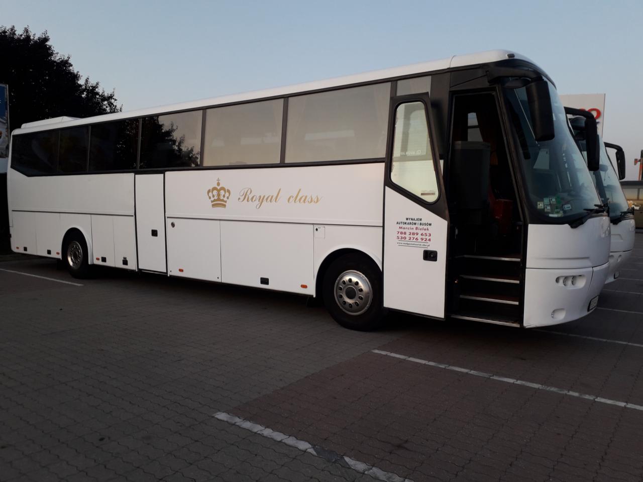 Biały autokar 'Royal Class' z otwartymi drzwiami, zaparkowany na kostce brukowej; widoczny numer telefonu firmy.