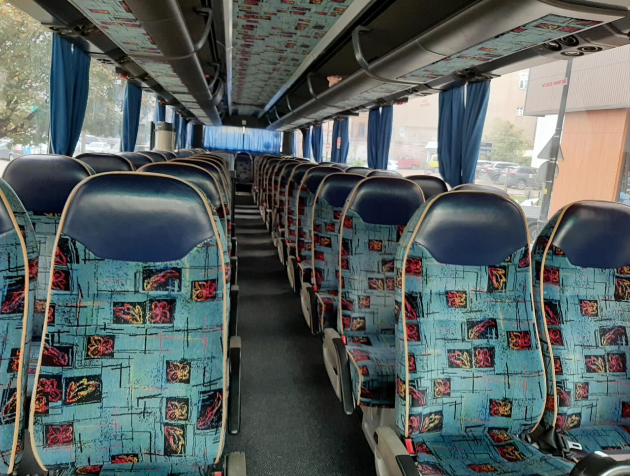 Wnętrze autobusu z rzędami niebiesko-turkusowych foteli w geometryczne wzory, zasłonięte niebieskie zasłony przy oknach, widok na ulicę z zaparkowanymi samochodami.