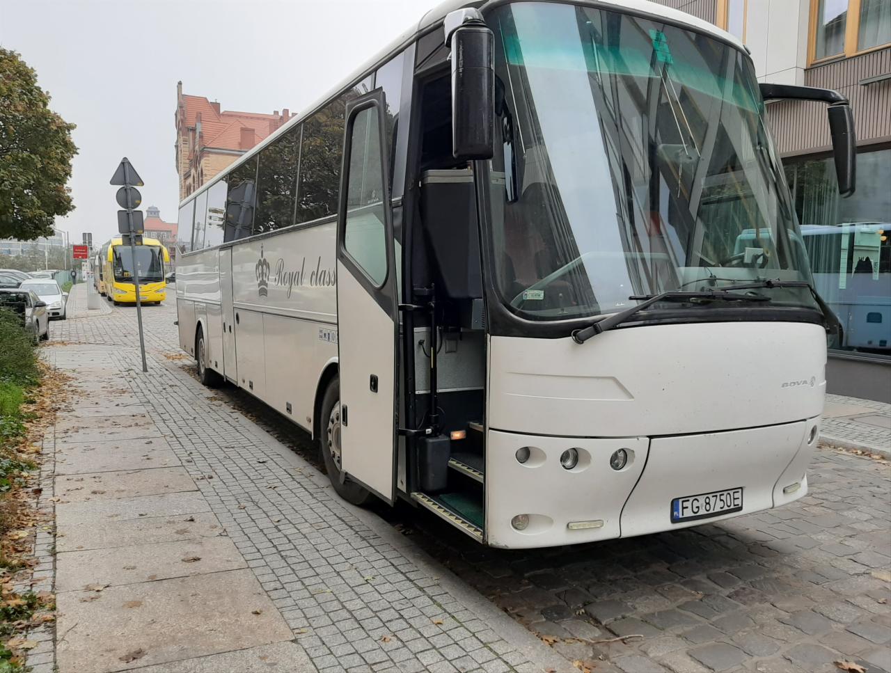 Biały autokar z otwartymi drzwiami na chodniku z kostki brukowej, widoczny napis 'Royal class' i numer rejestracyjny FG 8750E.