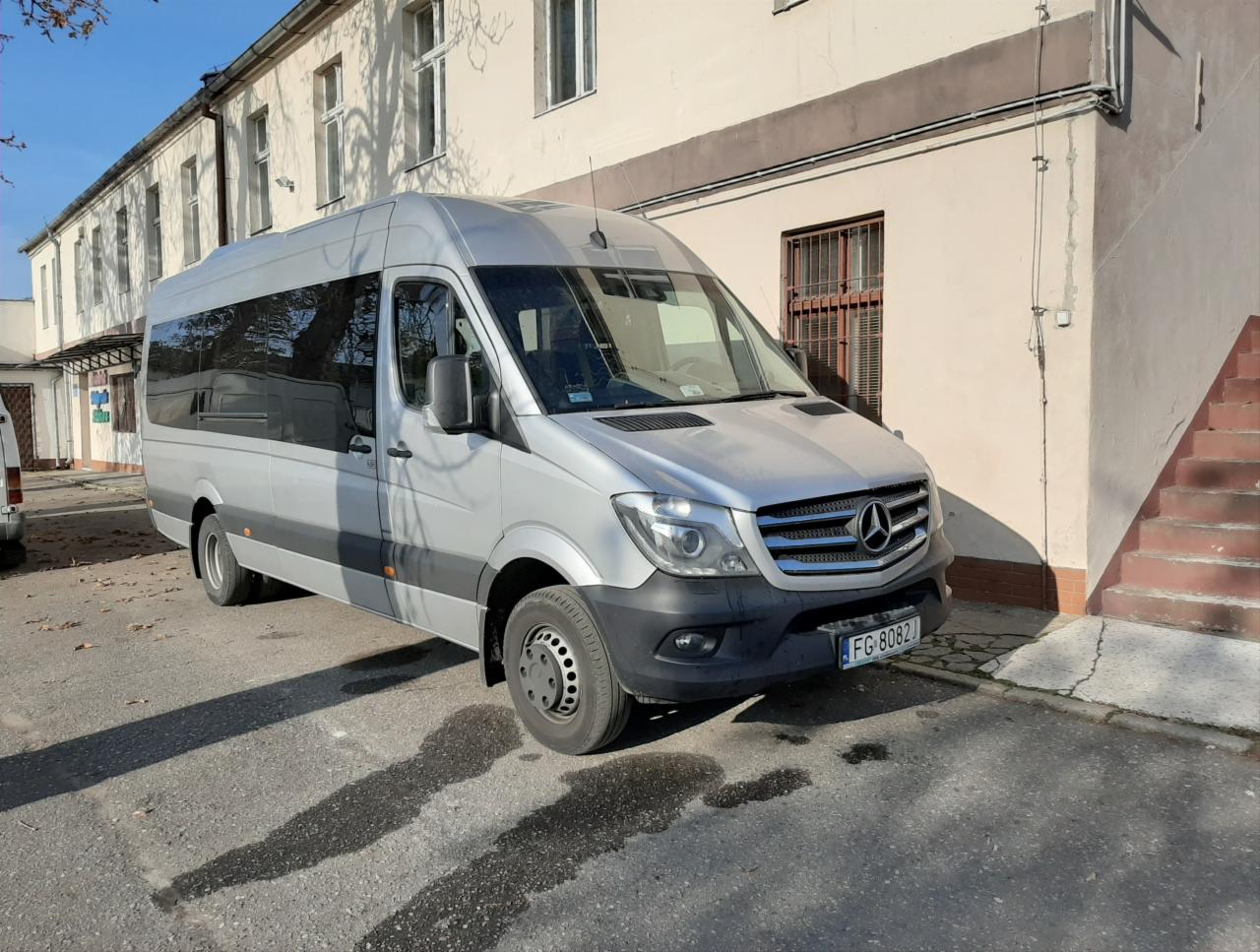 Srebrny bus Mercedes Sprinter z ciemnymi szybami, zaparkowany przed budynkiem z ceglanymi schodami. Widoczne tablice rejestracyjne FG 8082.