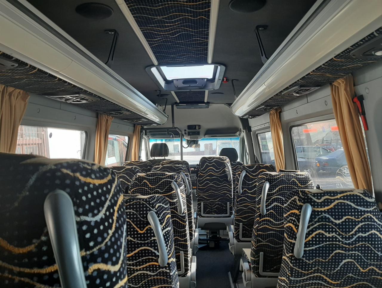 Wnętrze busa pasażerskiego z rzędami foteli w czarno-żółte wzory, widok z tyłu pojazdu, okna z zasłonkami, oświetlenie sufitowe.