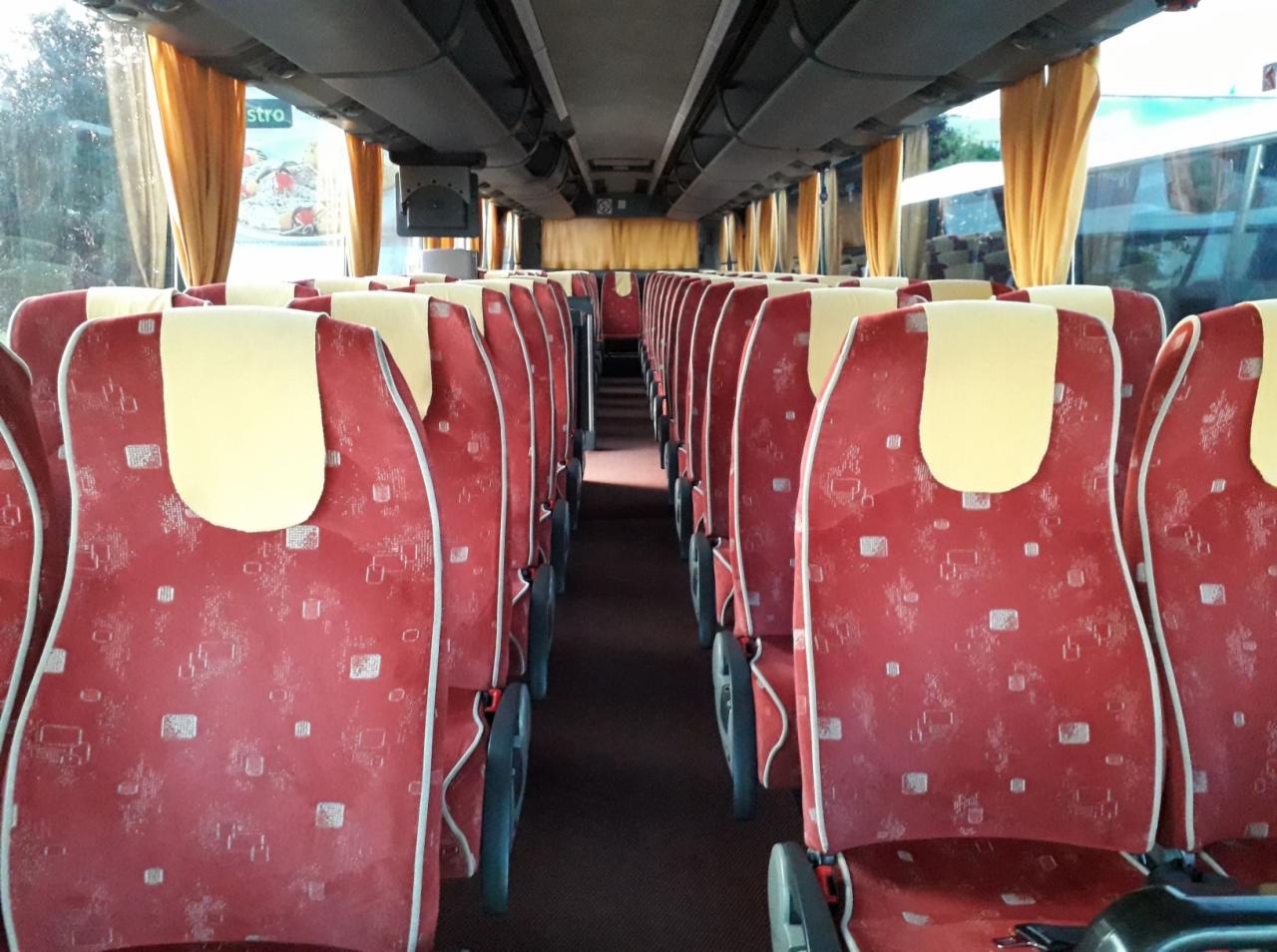 Wnętrze autobusu z czerwonymi fotelami w geometryczne wzory i żółtymi zagłówkami, widok z perspektywy pasażera w kierunku tyłu pojazdu.