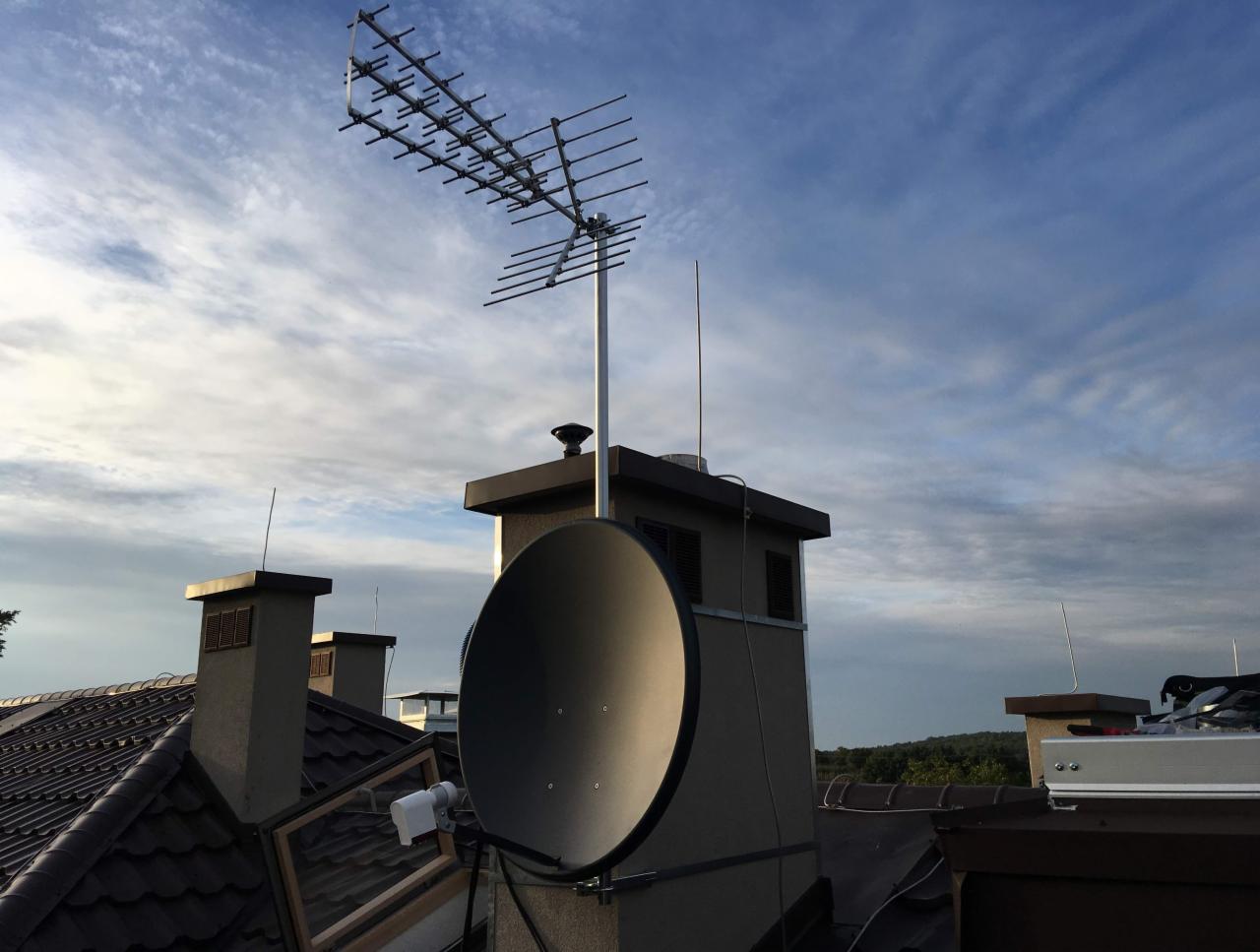 Antena telewizyjna Yagi-Uda i antena satelitarna zamontowane na kominie domu z brązową dachówką na tle błękitnego nieba z chmurami.