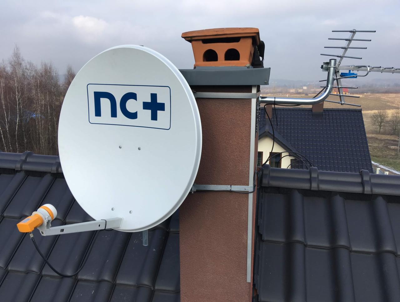 Antena satelitarna z logo nc+ i antena naziemna zamontowane na kominie domu z ciemną dachówką, widok z góry.