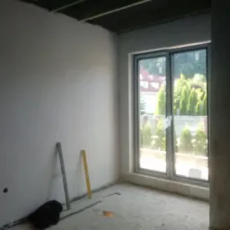 Pokój po tynkowaniu maszynowym, widoczne gładkie ściany, okno z widokiem na zieleń, poziomica i łatka tynkarska na podłodze.