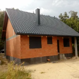 Surowy budynek jednorodzinny z czerwonej cegły z czarną dachówką i kominem, widoczne rolety zewnętrzne, otoczenie w stanie budowy, zarośla i drzewa w tle.