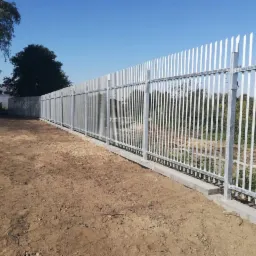 Długi, metalowy płot z ostrymi zakończeniami pionowych prętów, oddzielający teren z ziemią od drzew i zabudowań w tle, widziany z boku w słoneczny dzień.