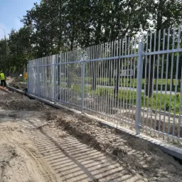 Montaż metalowego ogrodzenia panelowego na betonowej podmurówce, widoczne ślady prac ziemnych i pracownik w odblaskowej kamizelce w tle.