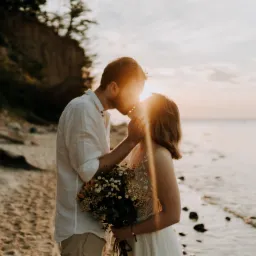 Para młoda całuje się na plaży o zachodzie słońca, mężczyzna w lnianej koszuli, kobieta w białej sukience trzyma bukiet polnych kwiatów, słońce przebija przez ich sylwetki.