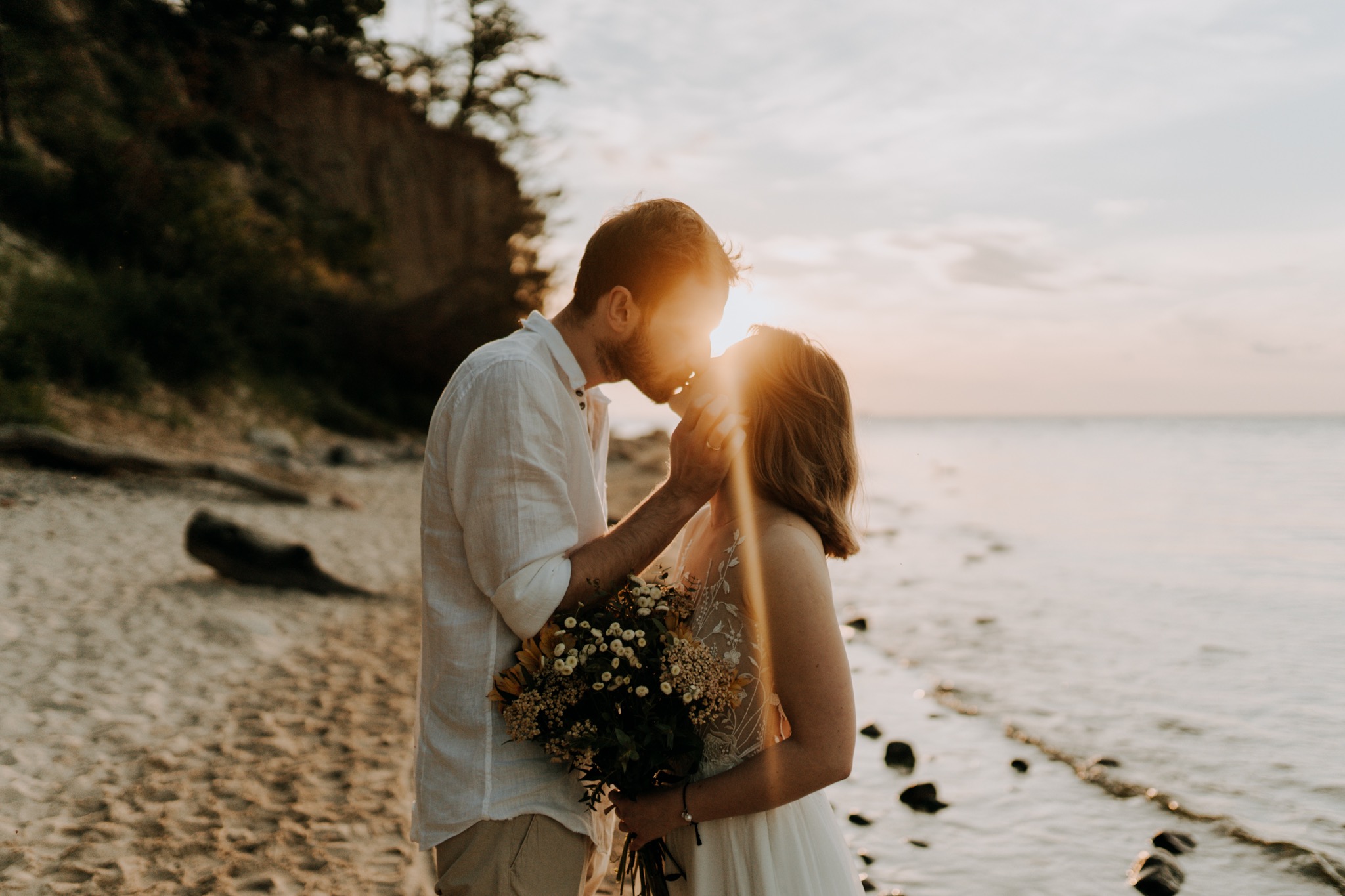 Para młoda całuje się na plaży o zachodzie słońca, mężczyzna w lnianej koszuli, kobieta w białej sukience trzyma bukiet polnych kwiatów, słońce przebija przez ich sylwetki.