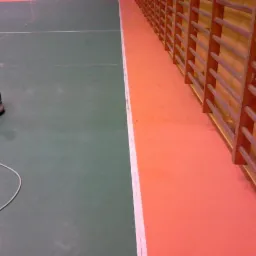 Maszyna do czyszczenia podłóg w hali sportowej, widoczna na zielonej podłodze obok pomarańczowego pasa z drabinkami gimnastycznymi.