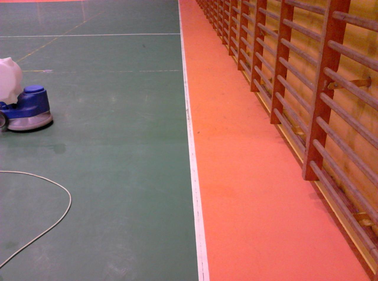 Maszyna do czyszczenia podłóg w hali sportowej, widoczna na zielonej podłodze obok pomarańczowego pasa z drabinkami gimnastycznymi.
